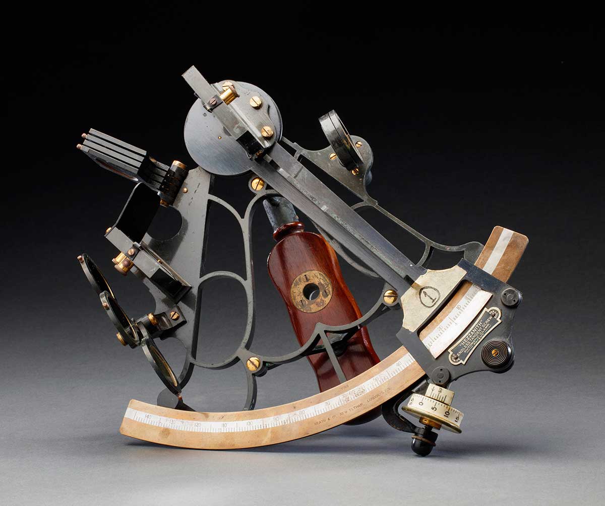 A marine model sextant made of black metal with brass fittings, in a wooden box, with various attachments.