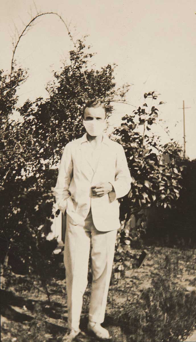 A sepia photograph of a man wearing a face mask and a white suit posing outside.