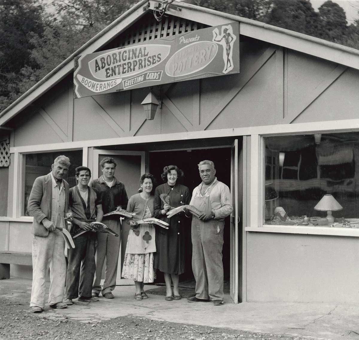 Black and white photograph of four men and two women standing outside a retail store called Aboriginal Enterprises. The signage above indicates the store's wares including boomerangs, greeting cards and handmade pottery.
