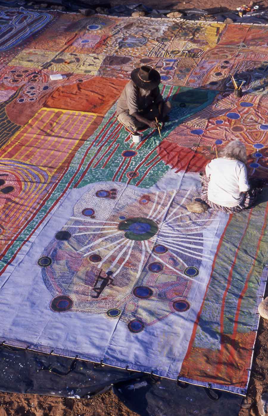 A man and a woman sitting on a large canvas to paint a section of the artwork.