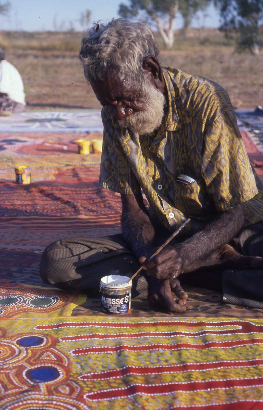 A man sitting on a large canvas with a paint brush, and white paint to working on the art piece.