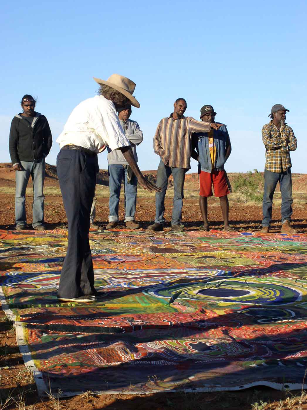 A man wearing a cowboy hat stands on a large colourful canvas in the desert, while several other people look on.