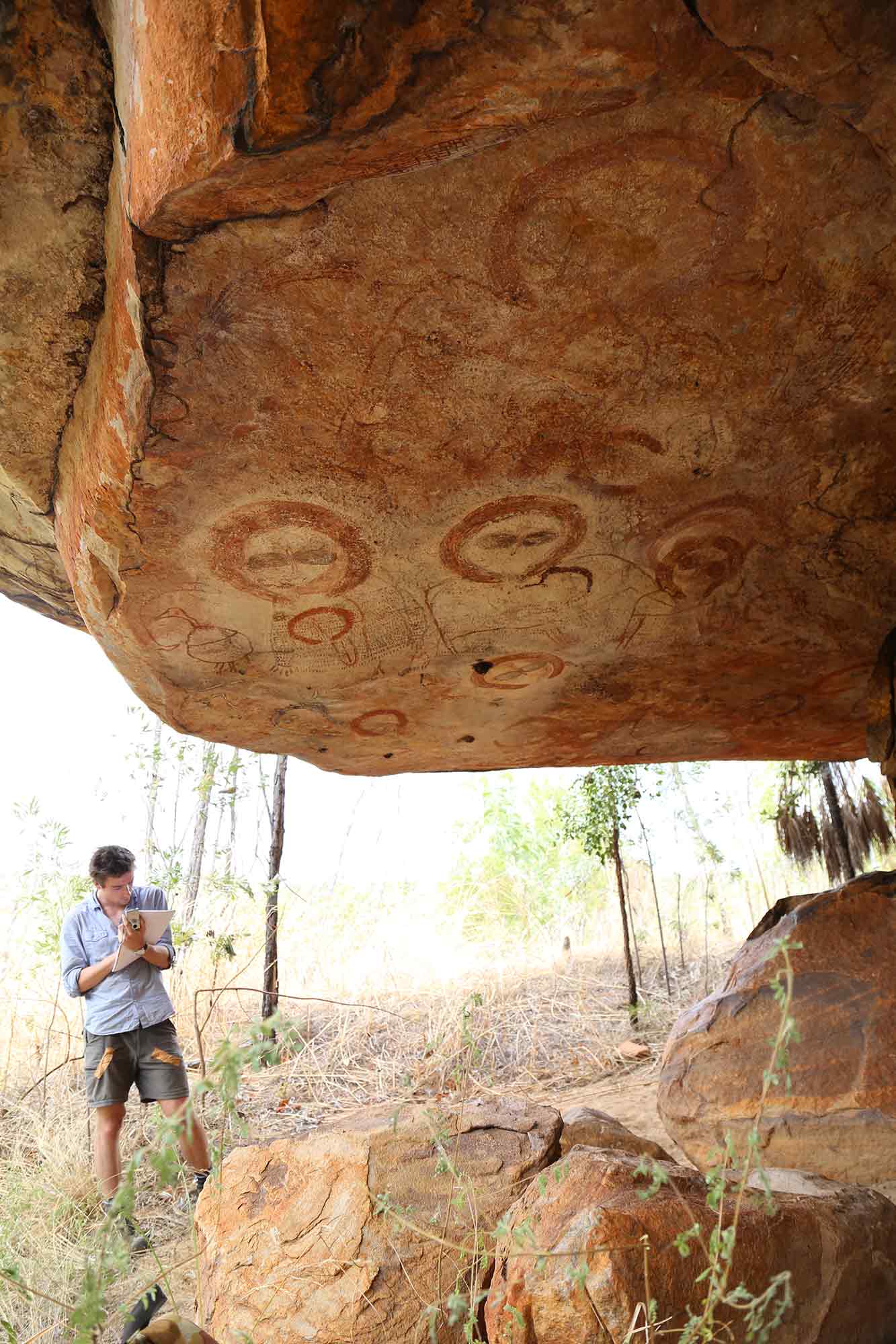 A man writes in a notebook at the entrance to a cave with rock art on the ceiling.