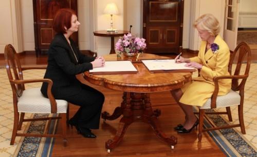 Julia Gillard is sitting at a table with Quentin Bryce who is signing an official document.