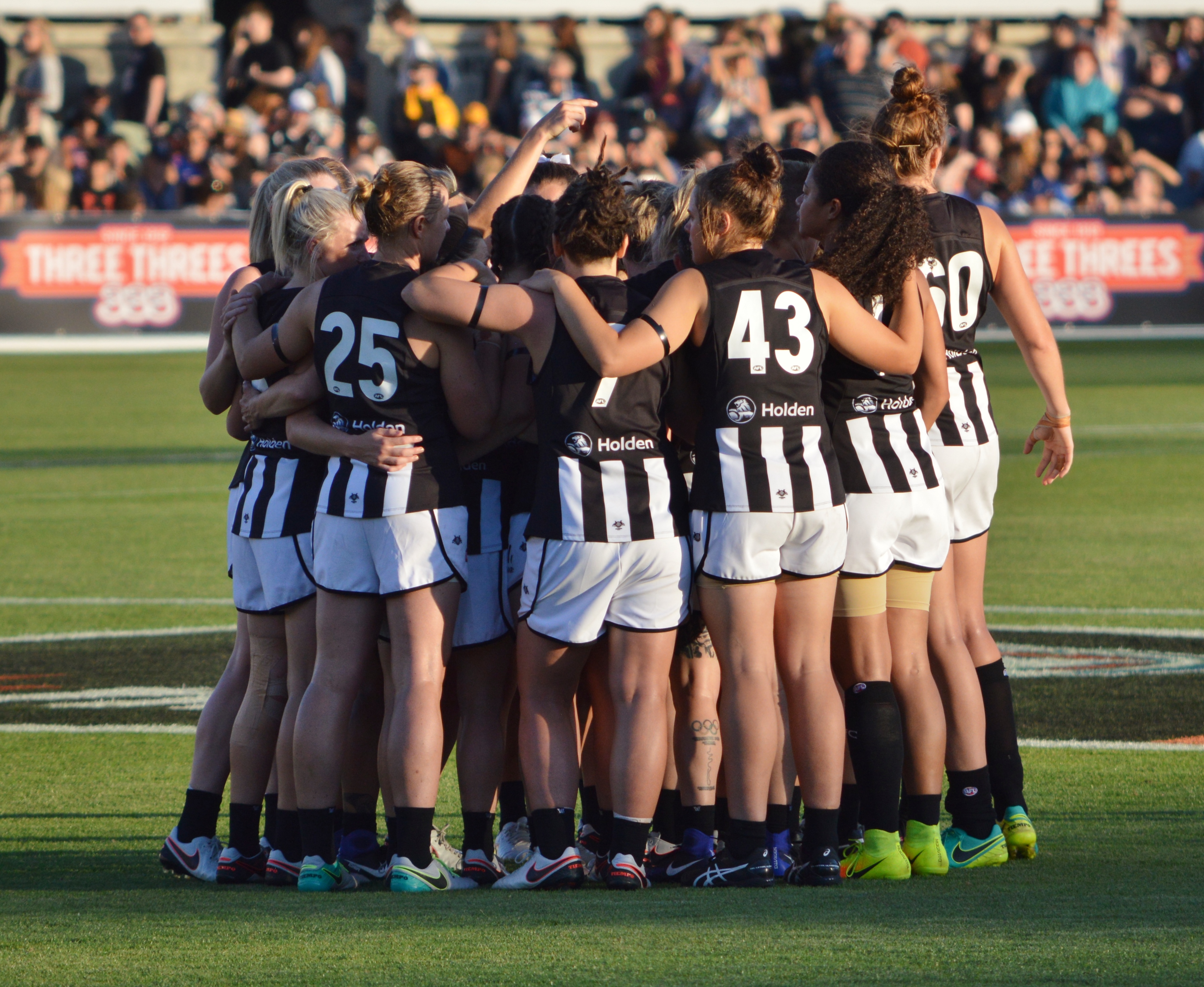 The Collingwood AFLW team huddled. The players are wearing black and white stripped Collingwood jerseys and white shorts. 