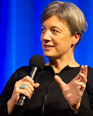 A headshot photo of a Michelle Simmons holding a microphone and looking to her right. Michelle has short grey hair, is wearing a black shirt and large blue ring.