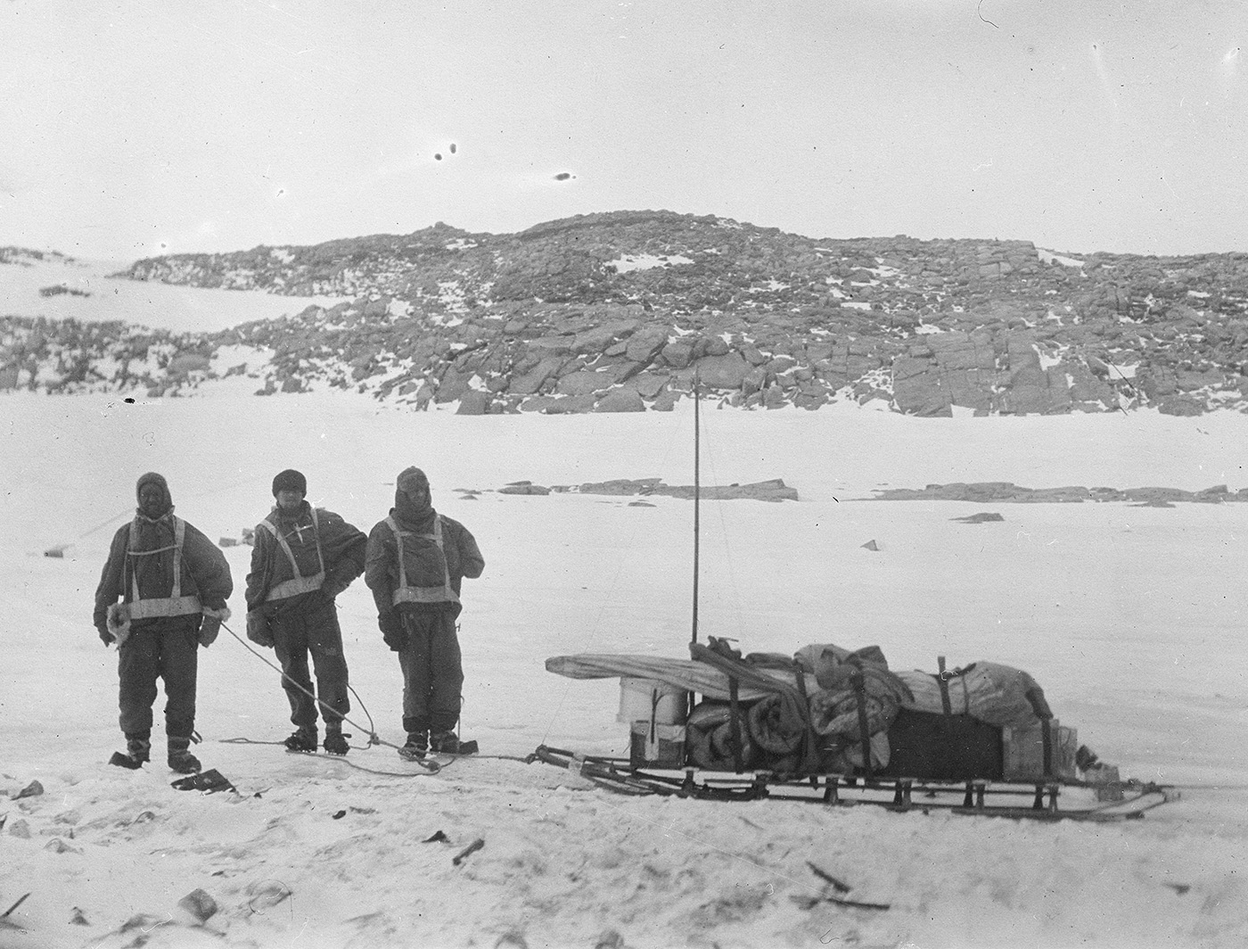 A black and white historical photograph where three men dressed in winter layers stand to the left, facing the camera. The men are hauling attached by harnesses to a large snow sled piled with supplies. Rocky hills in the background, snow in the foreground.