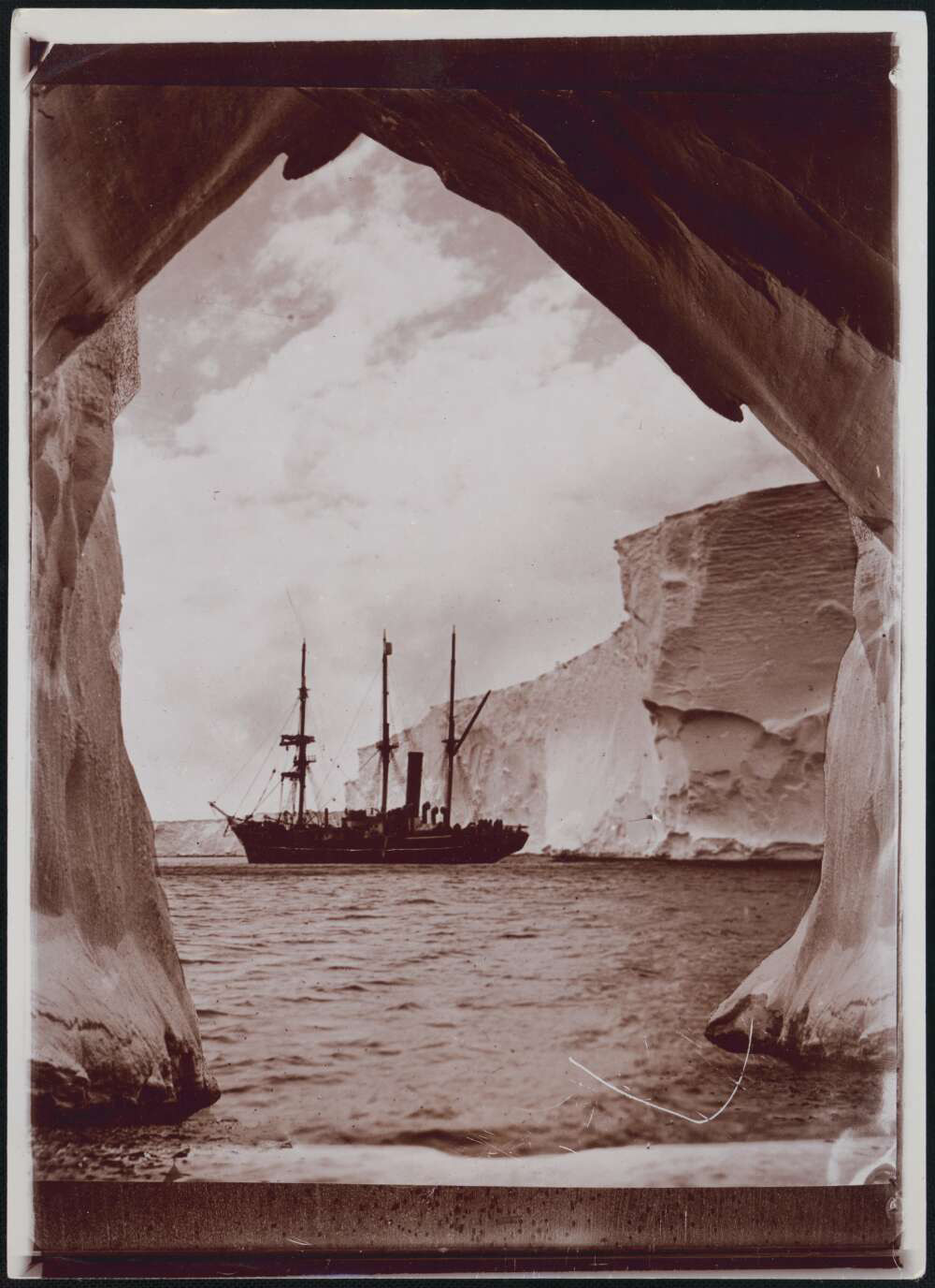 Photograph of a ship on water surrounded by icebergs, from the perspective of an iceberg archway.