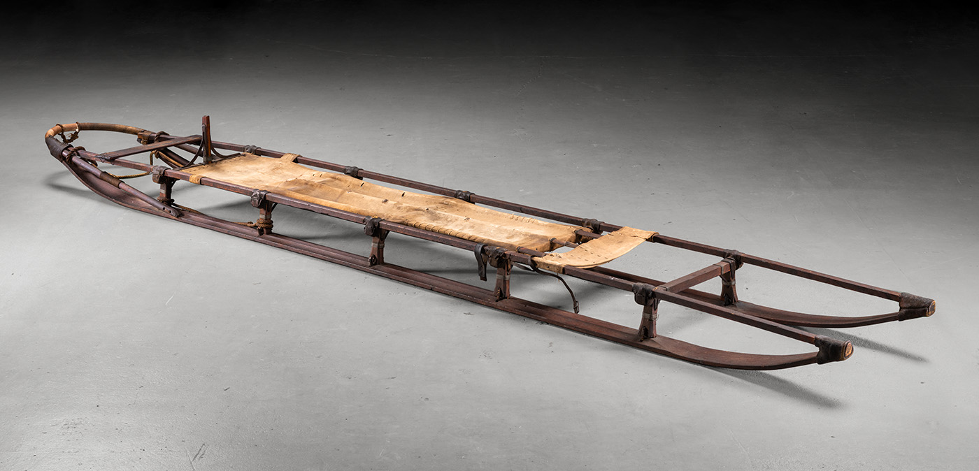 Alt text: A large brown wooden sled with rope tied to it, contrasted by a light grey background.