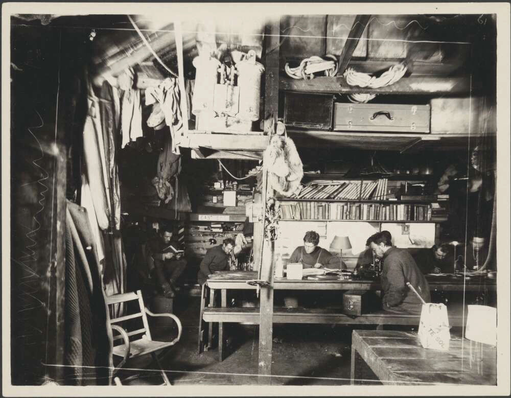 A black and white photograph shows an interior of a hut with six figures sitting around a wooden table in front of a wall with a large set of bookshelves.
