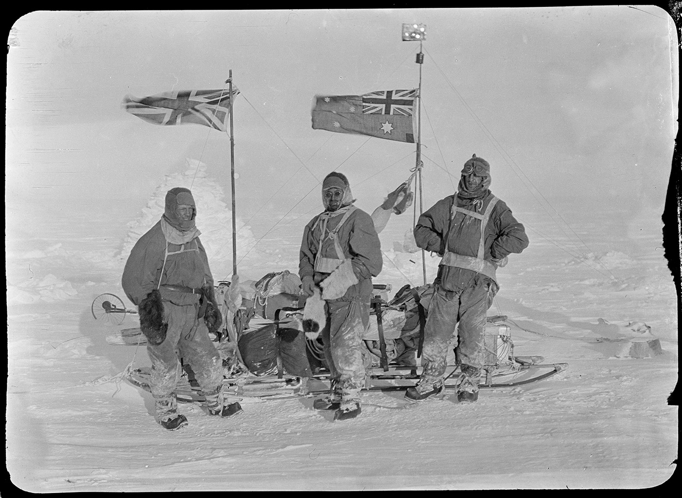 Three figures stand in front of a full sled, surrounded by snow. All figures are facing the camera, with both Australian and United Kingdom flags attached to the sled.
