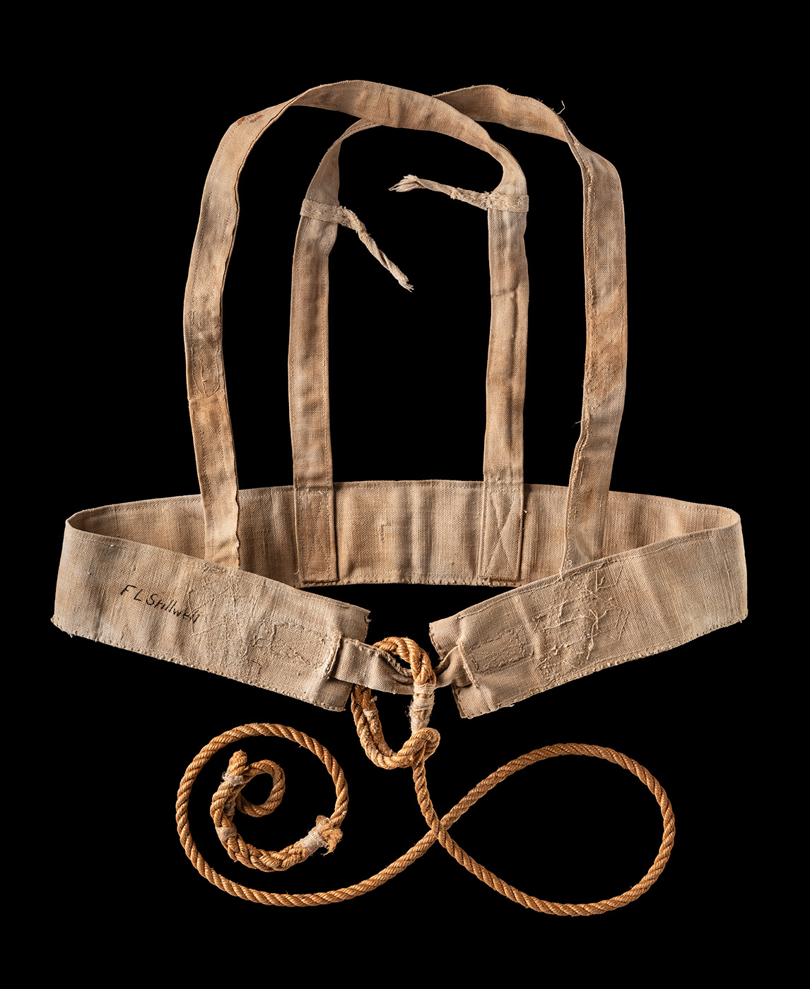 A light-coloured canvas harness is contrasted with a black background. There is a curled piece of rope attached to the harness, and the harness is signed 'F L Stillwell.' 