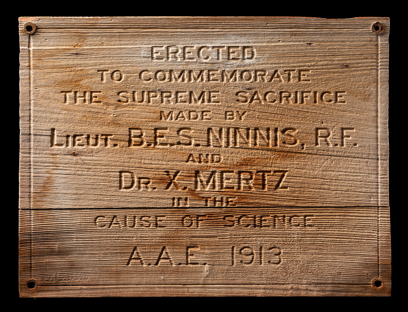Rectangular wooden sign with a nail in each corner. It reads: 'ERECTED TO COMMEMORATE THE SUPREME SACRIFICE MADE BY LIEUT. B. E. S. NINNIS, R. F. AND DR. X. METZ IN THE CAUSE OF SCIENCE A. A. E. 1913'.