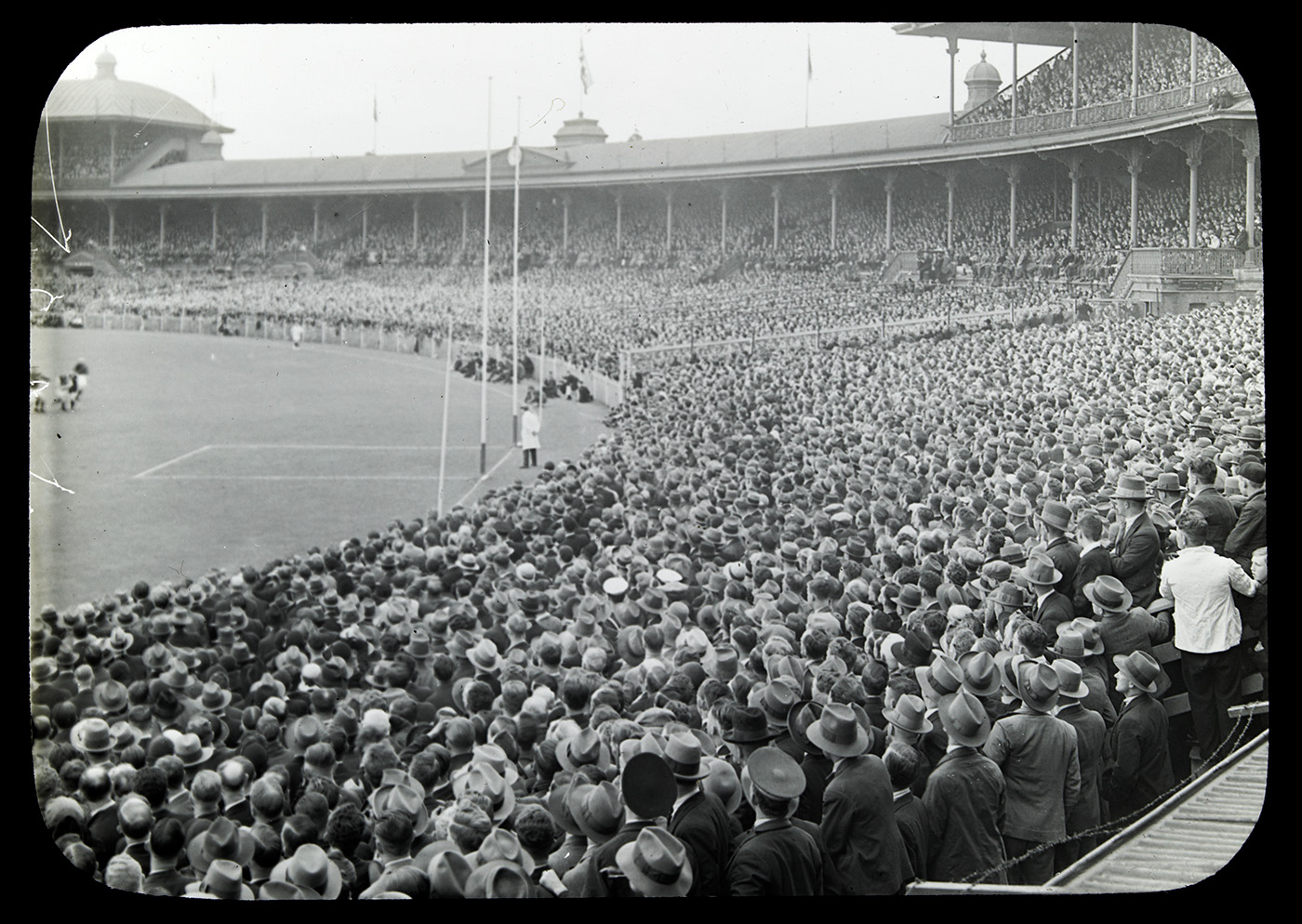 Black and white photograph showing a large crowd of people watching a game of Australian Rules football, with a man in white coat standing between goal posts at one end.