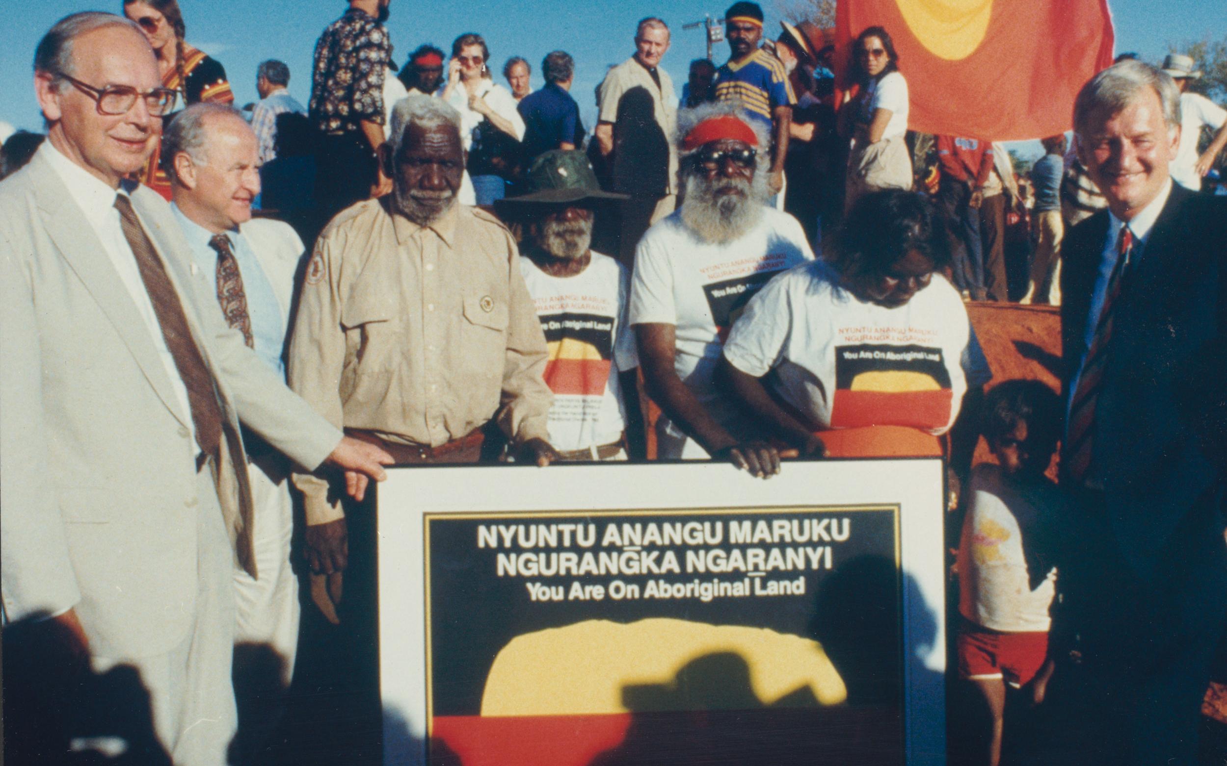 Traditional owners Mr Peter Bulla, Mr Peter Kanari, Mr Nipper Winmarti and his wife, Barbara Tjirkadu with Sir Ninian Stephen, Mr Holding and Mr Cohen (extreme right) and the special poster marking the handback of the title to Uluru-Kata Tjuta National Park.