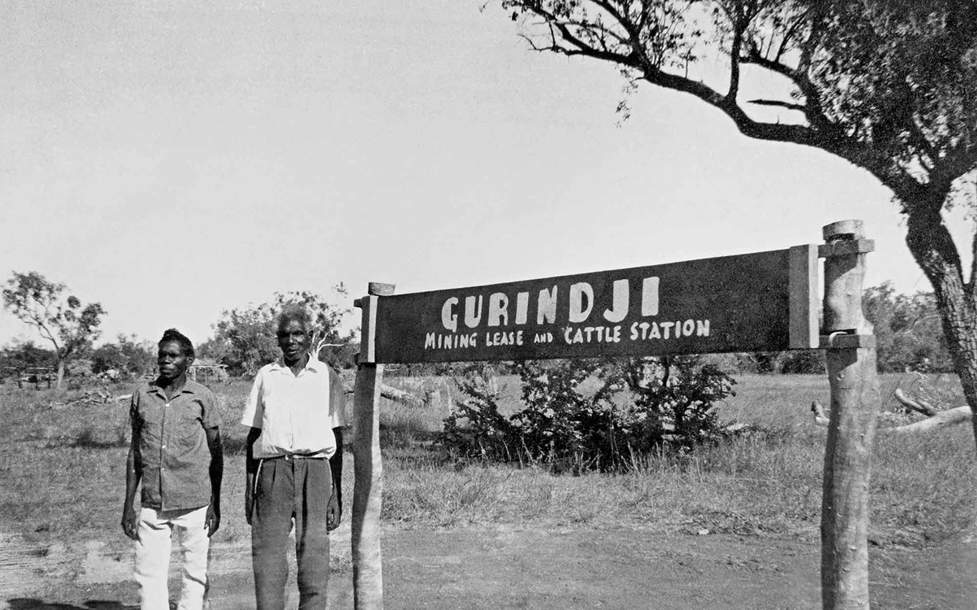 Black and white photo of two men beside a sign reading ‘Gurindji mining lease and cattle station’. 