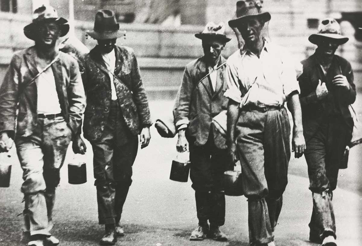 Black and white photograph of five men walking towards the camera dressed in shabby clothes. Most are carrying swags and billy cans.